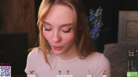 Video of a young, fair-skinned, blonde woman with long hair, wearing a white shirt with small animal prints, focusing intently on a smartphone. Background shows a dark room with a TV and a vase of blue flowers.