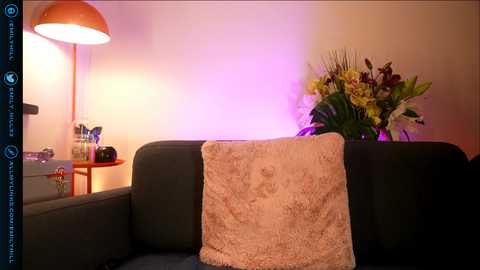 Video of a cozy living room with a dark sofa, a beige textured pillow, a vibrant floral arrangement, and a modern lamp emitting warm light.