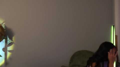 A video shows a woman with long black hair in a dimly lit room, sitting on a green cushion, holding her face. The background features a plain beige wall and a lamp with a yellow shade on the left.