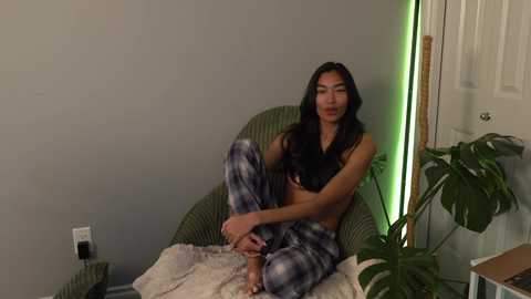Video of a slender, dark-haired woman in a tie-dye top and pants, sitting on a wicker chair in a dimly lit room with green and white walls.