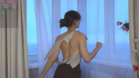 A video of a slender, fair-skinned woman with brown hair in a bun, wearing a revealing, backless, white top, and black pants, standing near a window with sheer curtains and a vase of flowers.