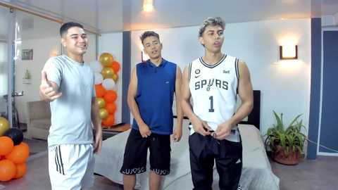 Video of three young Asian men in basketball gear, celebrating indoors with balloons and plants, smiling.