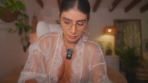 Video of a Latina woman with long black hair, wearing glasses and a sheer white lace robe, leaning forward, showing cleavage. Background includes wooden beams, potted plants, and a cozy indoor setting with warm lighting.