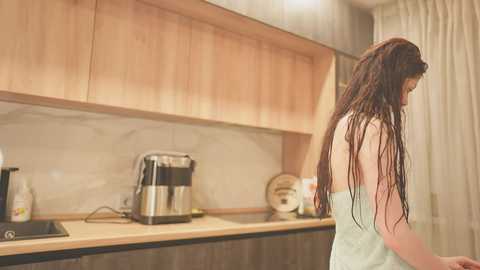 A video of a woman with long, wet, dark hair, wrapped in a towel, standing in a modern, minimalist kitchen with light wooden cabinets, stainless steel appliances, and beige walls.