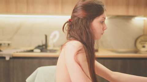 A video of a topless young woman with long brown hair, seated in a modern kitchen with wooden cabinets, stainless steel appliances, and a beige backsplash.