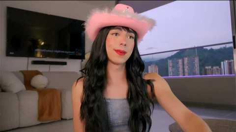 Video of a fair-skinned person with long black hair, wearing a pink cowboy hat, red lipstick, and a denim top, sitting in a modern living room with a TV and city skyline view.