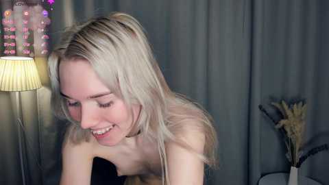 Video of a smiling, topless, fair-skinned blonde woman with straight hair, wearing a choker, in a dimly lit room with a lamp, gray curtains, and decorative pampas grass.