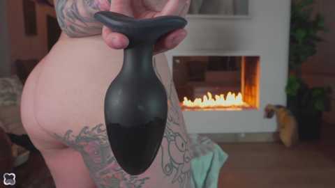 Video of a nude tattooed person holding a black anal plug, standing beside a lit fireplace with a green couch in the background.