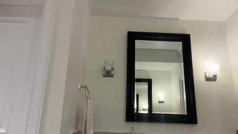 Video of a minimalist bathroom with a large black-framed mirror, two wall sconces, and a white towel rack. The walls are light gray, and the room is clean and modern.