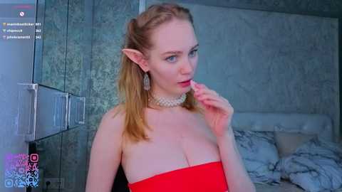 Video of a pale-skinned, blonde-haired, elf-like woman with pointed ears, wearing a strapless red dress and pearl necklace, biting her finger in a modern, minimalistic bedroom.