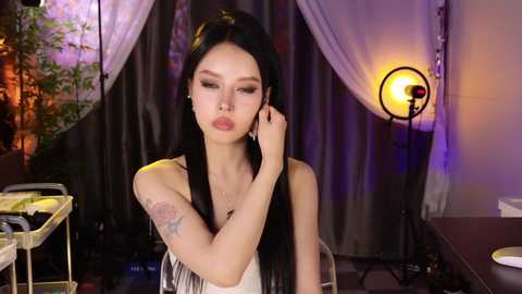Video of an East Asian woman with long black hair, light skin, and a tattoo on her arm, wearing a sleeveless top, in a dimly lit studio with hanging lights and greenery.