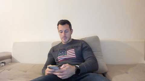 A video of a man sitting on a beige sofa, wearing a grey sweatshirt with an American flag design and holding a smartphone. The background is plain white.
