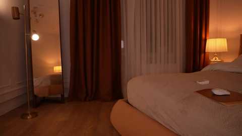Video of a warmly lit, modern bedroom with beige and brown tones. A large bed with a beige cover is against a backdrop of sheer white curtains and dark brown drapes. A standing lamp and a mirror are positioned on the left side.
