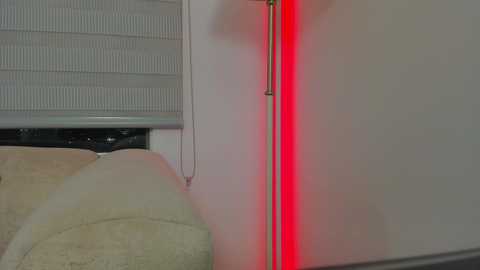 Video of a modern living room corner with a beige couch, a gray Venetian blind, a sleek silver floor lamp emitting red light, and a minimalist white wall, creating a contemporary, cozy atmosphere.
