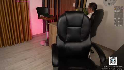 Video of a modern office with a black leather chair facing a desk; a man in a white shirt is seated. Background includes a wooden desk, bar stools, and dark curtains.