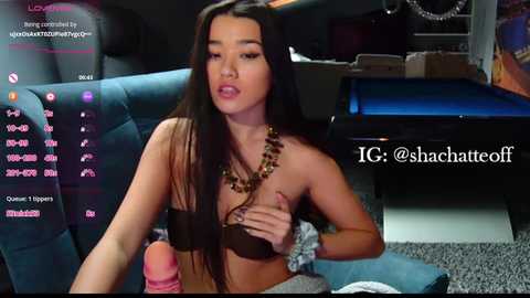 A video shows a topless Asian woman with long black hair, wearing a necklace, and a large, erect pink dildo in a dimly lit room with a blue couch and gaming setup.