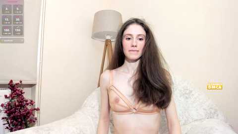 Video of a slender, Caucasian woman with long brown hair, wearing a revealing, peach-colored lingerie bra, sitting on a bed with white linens, in a softly lit, beige-toned room.