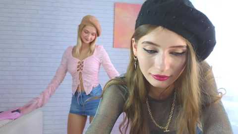 Video of a blonde woman in a pink lace top and denim shorts, smiling, and a brunette woman in a black beret and sheer top, looking down, in a brightly lit room with white brick walls.