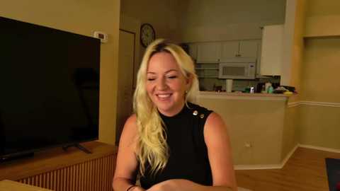 Video of a smiling blonde woman with long hair, wearing a black sleeveless top, seated in a modern, dimly-lit kitchen with white cabinets, wooden floors, and a large flat-screen TV on the left.