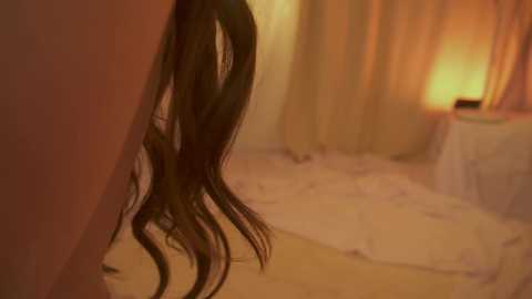 A close-up video of a woman's dark, wavy hair cascading over her shoulder in a dimly lit, softly colored bedroom setting with beige curtains and a warm, ambient light.