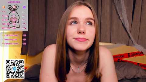 A video of a young, fair-skinned woman with long, straight, light brown hair, wearing a silver necklace, smiling slightly. She is topless, sitting on a bed with grey and red sheets. The background has brown curtains and a QR code.