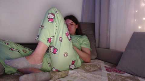 Video of a woman with light skin and dark hair, wearing light green pajamas with Hello Kitty patterns, lying on a bed with a quilted bedspread, in a dimly lit room.
