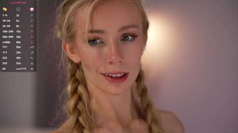 Video of a smiling, fair-skinned blonde woman with braided hair, wearing a strapless top. Background is blurred, showing a dimly lit room.