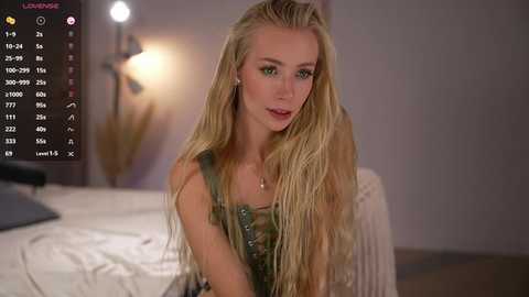 Video of a fair-skinned, blonde-haired woman in a green dress, seated on a bed in a dimly-lit room. Background includes a bedside table with a lamp and a vase of dried flowers.