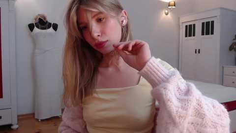 Video of a young woman with long blonde hair, wearing a yellow tank top and a knitted pink cardigan, posing in a brightly lit, modern bedroom with white furniture and a mannequin.