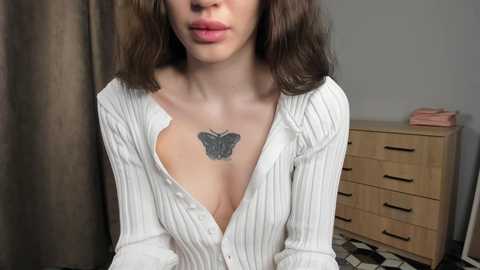 A video of a young woman with fair skin and wavy brown hair, wearing a white, ribbed knit cardigan that reveals a butterfly tattoo on her chest, sitting in a bedroom with a wooden dresser and checkered floor.