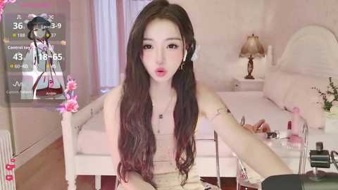 Video of a young Asian woman with long dark hair, wearing a beige off-shoulder dress, sitting in a cozy, pink-themed bedroom with a white bed, nightstand, and floral decor.