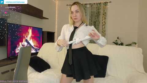 Video of a blonde woman in a white blouse and black skirt, adjusting her tie in a modern living room with a TV, fireplace, and fairy lights.