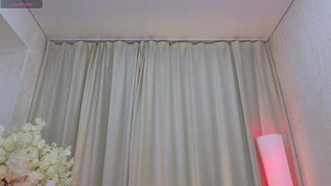 Video of a room featuring a white curtain that drapes over a window, partially covering a white floral arrangement. A tall, cylindrical white lamp with a red base is visible on the right.