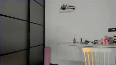 Video of a minimalist, modern bathroom with a white sink, black shelving unit, and a wooden chair.