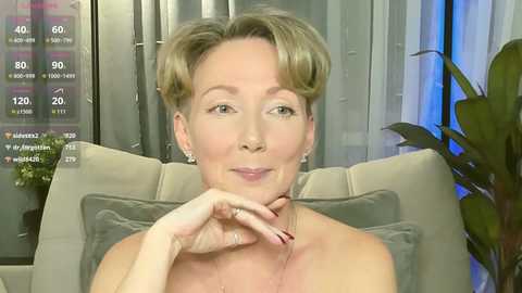 Video of a smiling, light-skinned woman with short blonde hair, wearing a gray dress, seated on a beige cushioned chair with a green plant in the background.