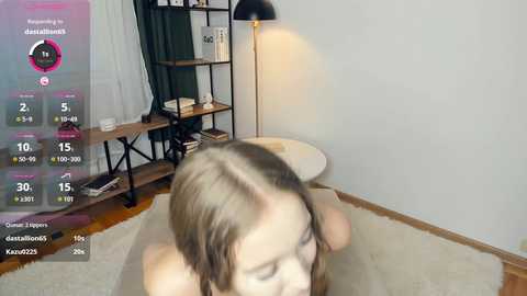 A video of a young woman with light skin and long brown hair, wearing a beige top, sitting on a white rug in a modern, minimalist room with wooden floors, a black lamp, and a bookshelf.