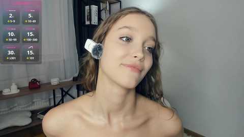 Video of a young Caucasian woman with light skin, long brown hair, and white headphones on her ears, applying makeup in a dimly lit room with a white wall and shelf.
