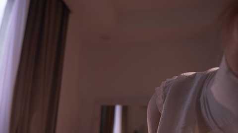 A dimly lit, close-up video of a person's upper body in a light-colored shirt, standing in a room with beige walls and dark curtains. The image is blurry, emphasizing soft shadows and a muted color palette.