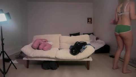 Video of a woman in green shorts and bra, standing in a dimly lit, cluttered bedroom with a white sofa, pink pillows, and a tripod-mounted camera.