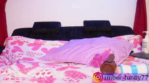 Video of a messy, pink-themed bedroom with a dark bed, pink sheets, and scattered sex toys, including a dildo and bottle of lube.