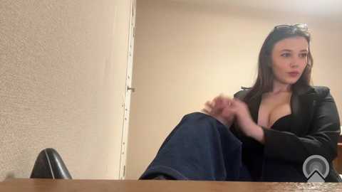 Video of a young woman with long dark hair, wearing a black blazer and low-cut top, adjusting her jeans while seated on a wooden desk in a beige-walled room.