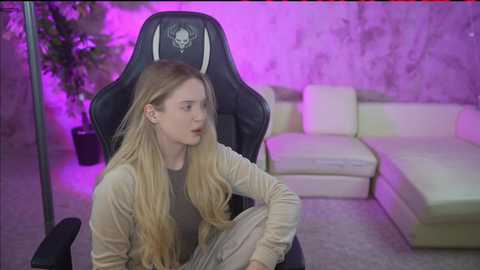 A video of a young, pale-skinned woman with long blonde hair, sitting in a black gaming chair, wearing a beige cardigan, looking distressed, in a modern, dimly-lit room with purple lighting, a white sectional couch, and a potted plant.