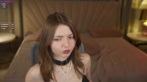 Video of a young woman with long brown hair, wearing a black choker and a black top, sitting on a bed in a dimly lit room.