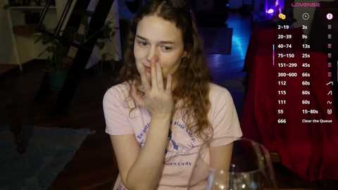 A video of a young woman with long, curly brown hair, wearing a pink T-shirt, covering her mouth in a dimly-lit living room, with a video feed overlay.