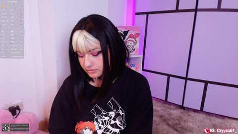A video of a young woman with a pale complexion, black hair with white streaks, and a nose ring, wearing a black graphic tee, sitting on a plush carpet in a room with pastel purple walls and a decorative screen.