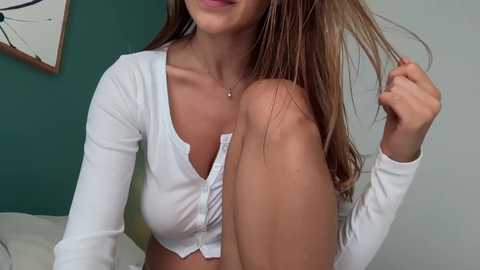 Video of a young woman with long, straight brown hair, wearing a white, long-sleeve, button-down crop top, sitting on a bed with white sheets.