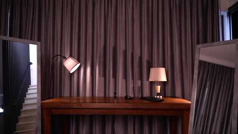 Video of a modern bedroom with a wooden desk, two lamps, and grey curtains; one lamp has a black metal stand, and the other has a white shade. The background includes a glimpse of a staircase.