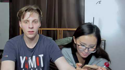 Video of a young man with short, light brown hair wearing a \"YOMOT\" t-shirt, and a young woman with long, dark hair and glasses, both seated on a bed, smiling, in a cozy, dimly lit room.