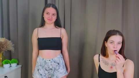 Video of two young women with fair skin and straight brown hair, one in a black crop top and floral shorts, the other in a black tank top, posing seductively.