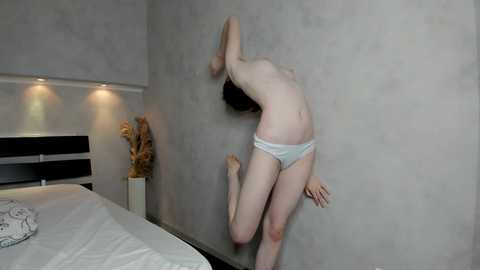 Video of a slender, topless woman with pale skin, wearing only white panties, jumping against a textured gray wall in a modern bedroom.